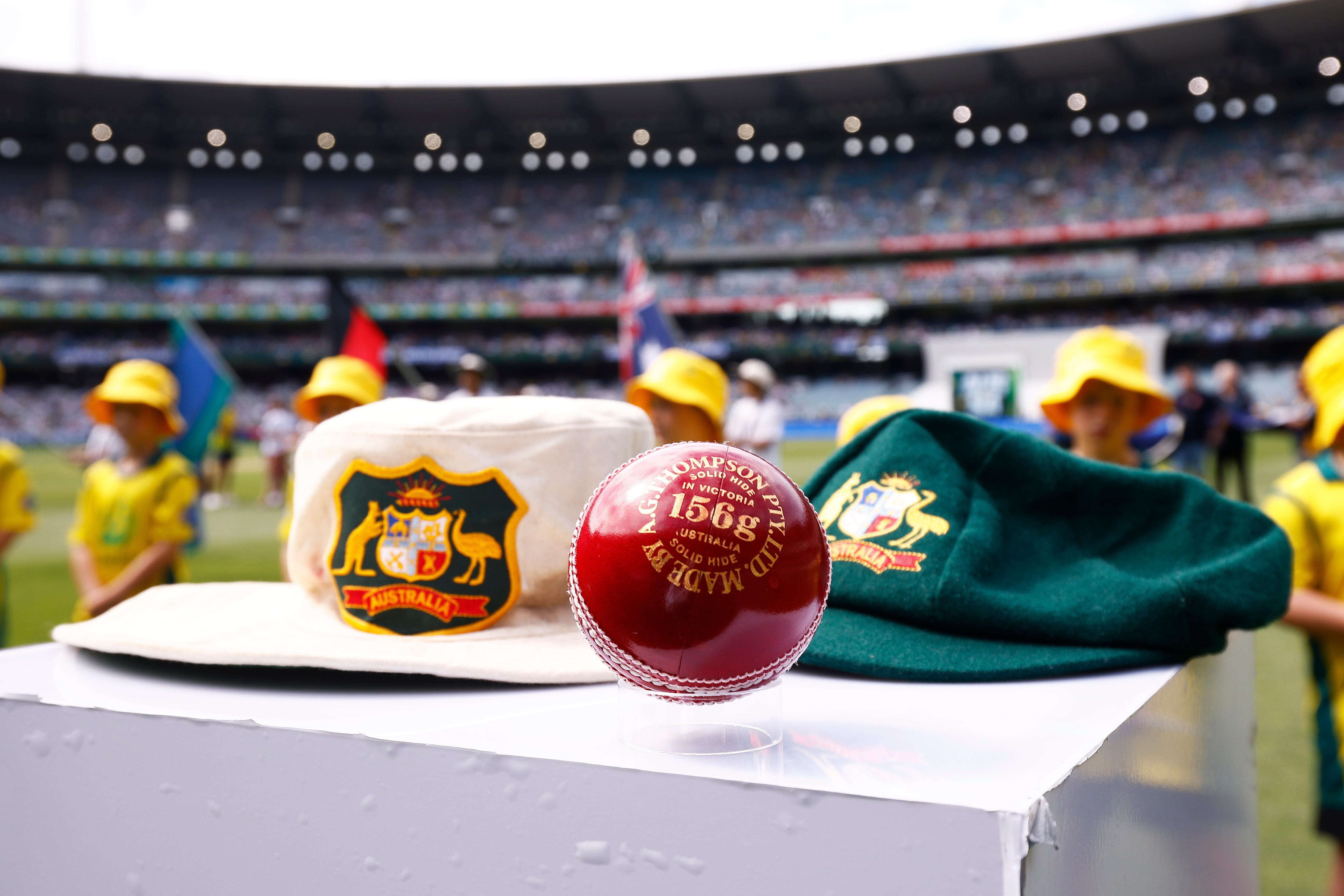 A close-up photo of a red cricket ball with a white floppy hat and baggy green in the background