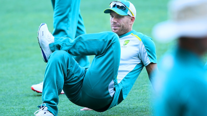 David Warner stretches out his glutes on the ground at training