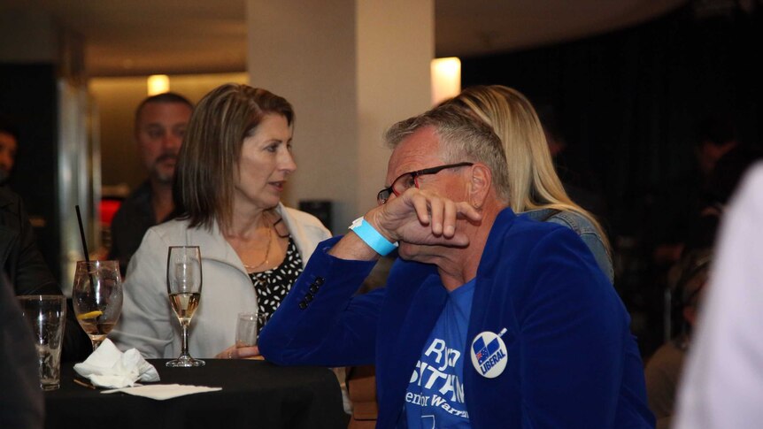 A man sits at a bench in a function room with his hand over his face.