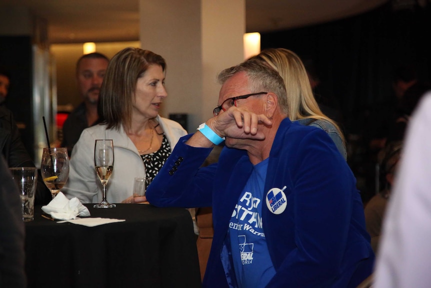 A man sits at a bench in a function room with his hand over his face.