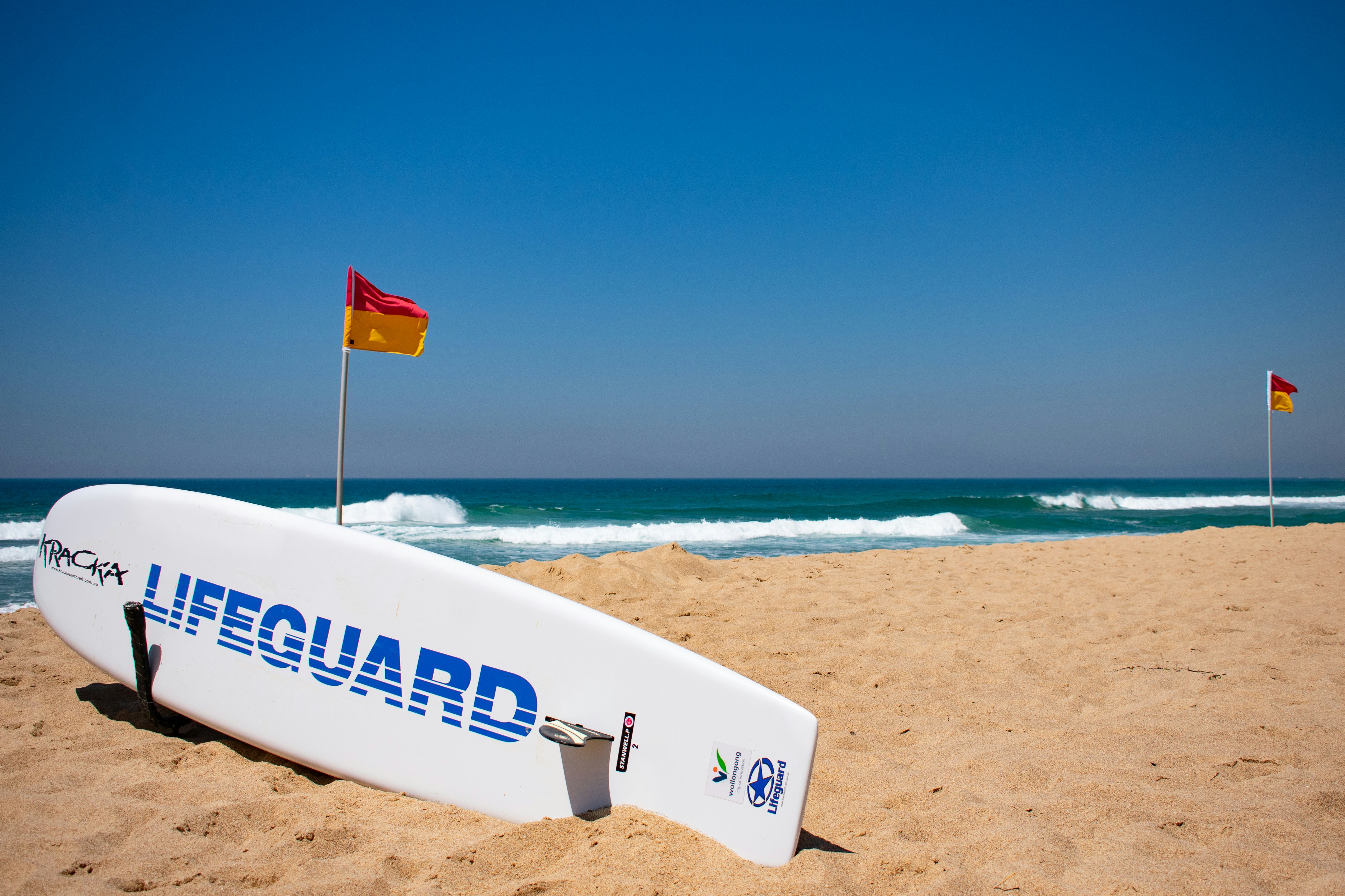 Beach with two red and yellow flags poled down on either ends with a lifeguard surfboard in the foreground