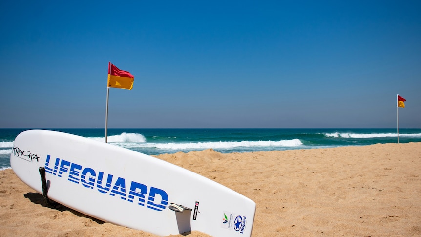 Beach with two red and yellow flags poled down on either ends with a lifeguard surfboard in the foreground