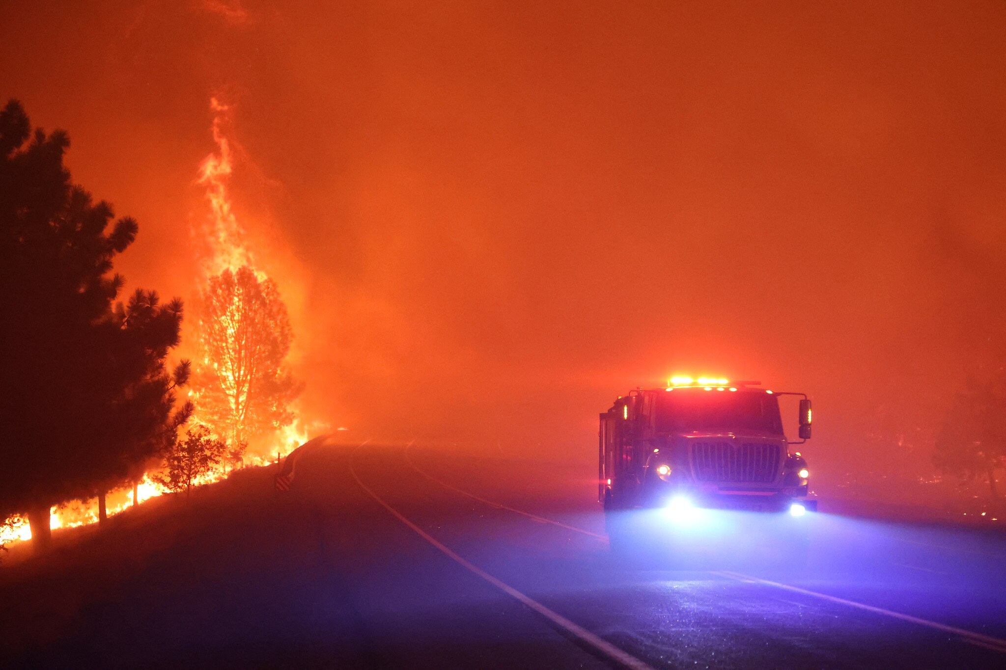 A fire truck drives through haze on a road alongside a burning forest 