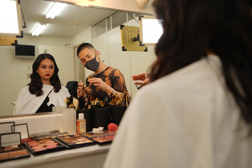 A man in a face mask holding a makeup brush looks in the mirror at a young woman sitting in a chair