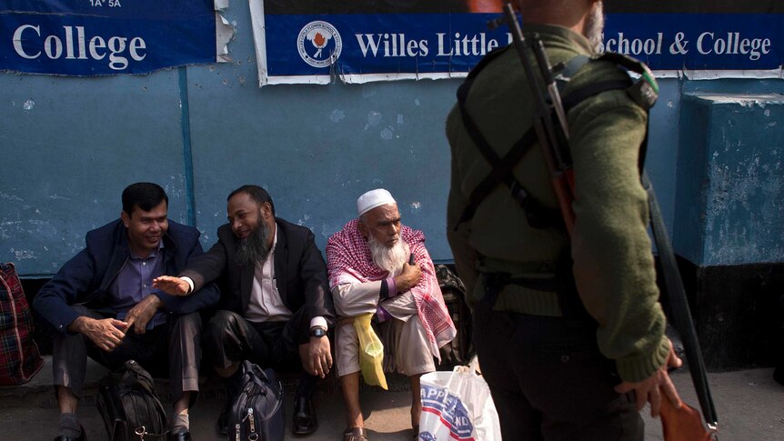 Bangladeshi men sit on the ground outside a polling station while a security official walks past with a rifle.
