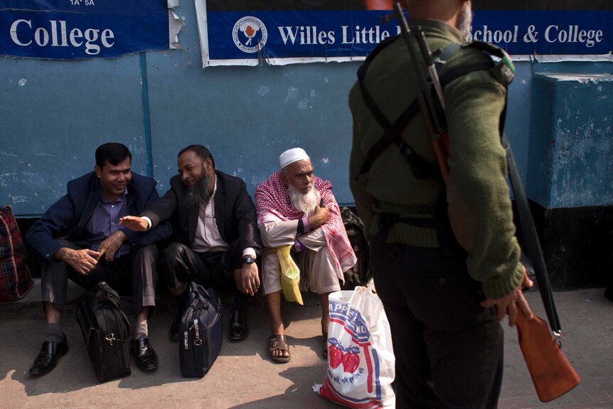 Bangladeshi men sit on the ground outside a polling station while a security official walks past with a rifle.