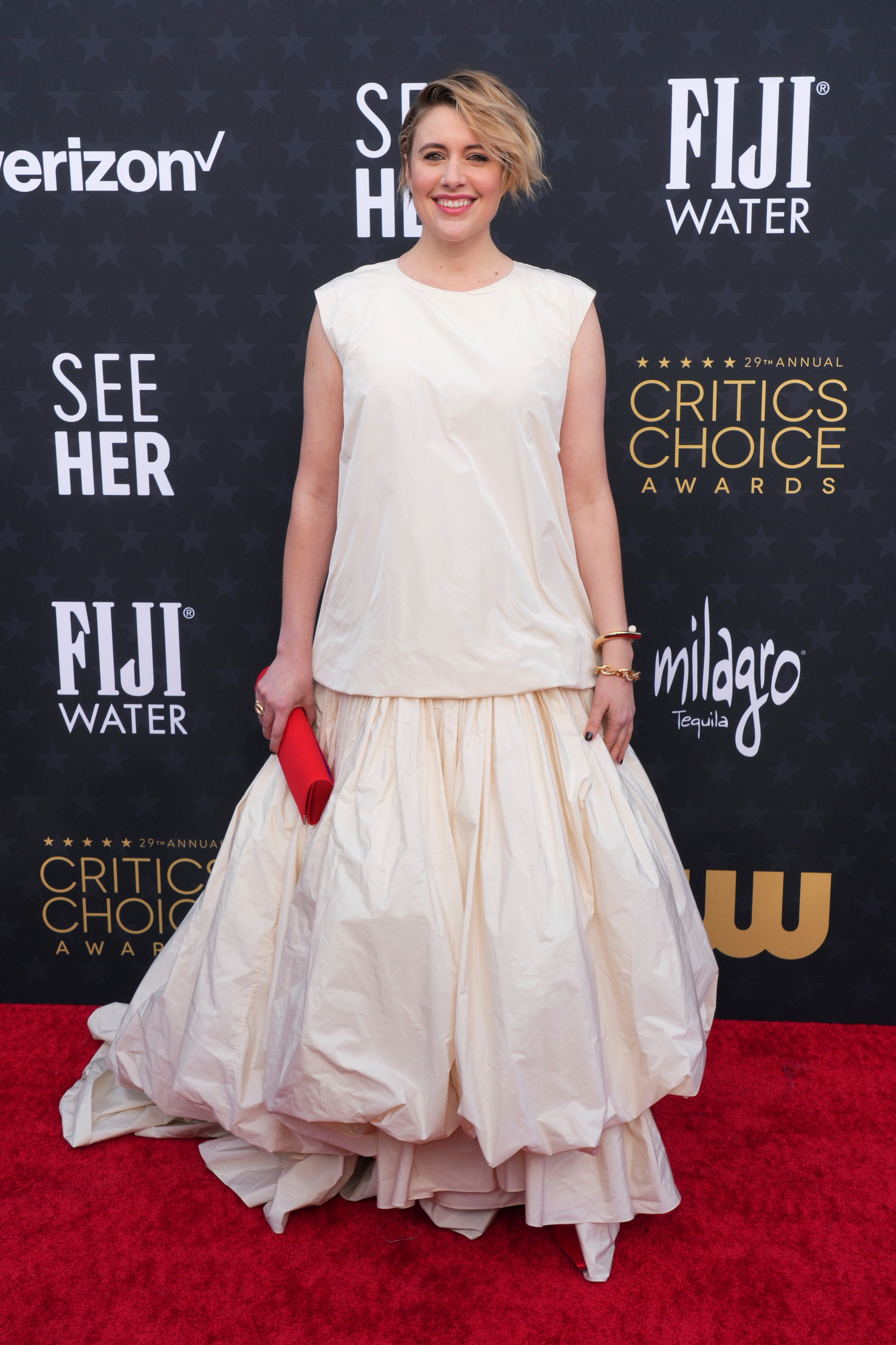 Greta Gerwig wearing a baggy white dress with sheet-like ruching and a bright red clutch and shoes 