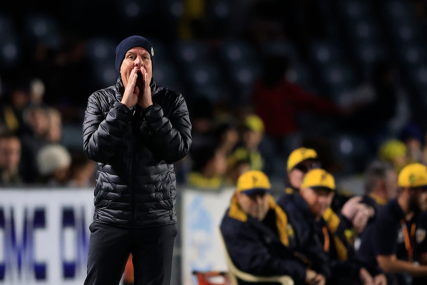 A man wearing a puffer jacket shouts on the sidelines of a sport event. 