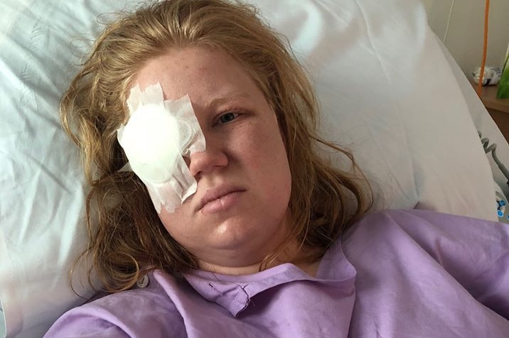 Rebecca Austin with looks at the camera with a cotton circle taped onto her face over her eye wearing a purple hospital gown.