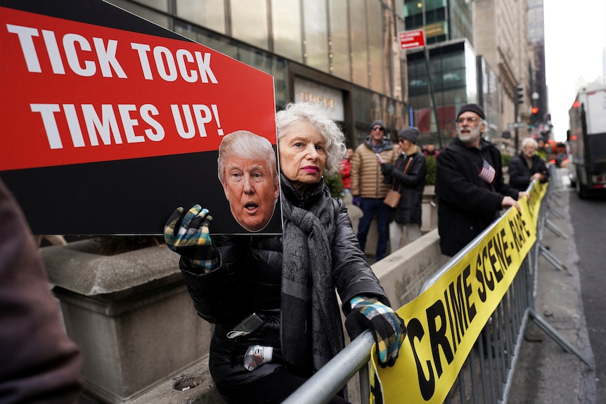 A woman holds a sign with a picture of Donald Trump on it which says 'tick tock times up'. 