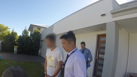 A man with his face blurred is led out of a house by police