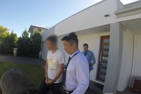 A man with his face blurred is led out of a house by police