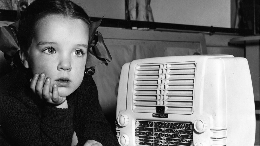 A black and white photo of a child with braids listening intenly next to a radio 