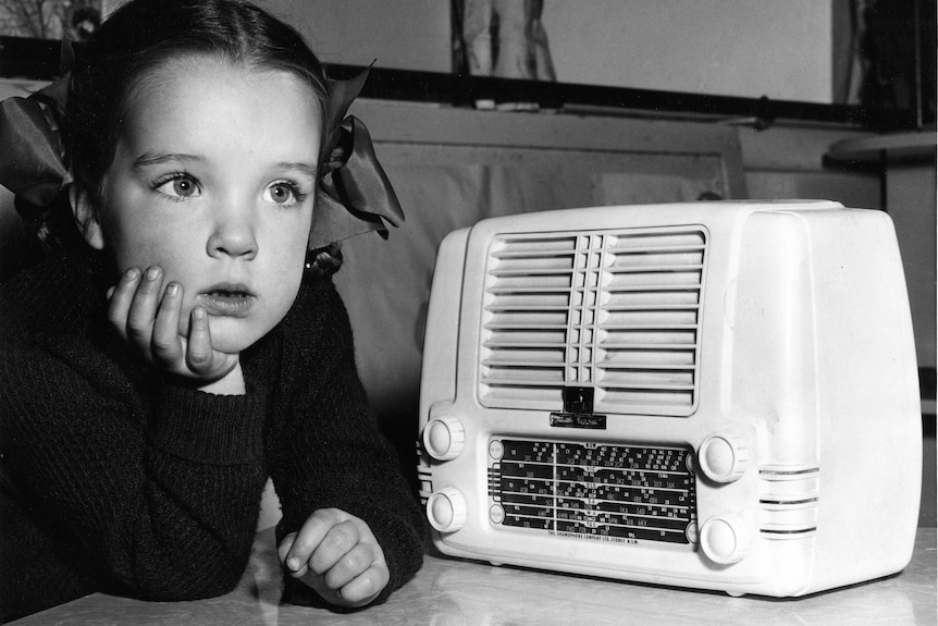 A girl listens intently next to an old-fashioned radio set.