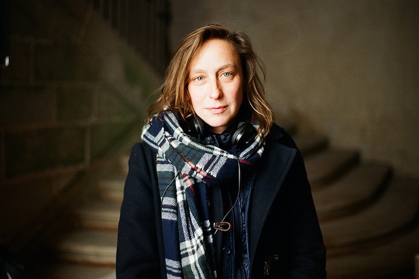 A man with shoulder length brown hair, headphones around neck, scarf and jacket stands in dim room in front of stairs.