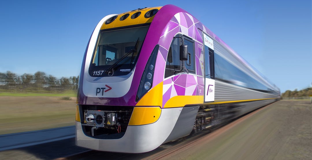 A purple, yellow and grey train moves along railway tracks in a rural setting.