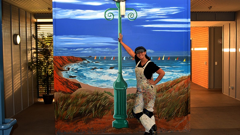 Sharron Tancred leans against a mural of a ocean scene that features a lamp post.