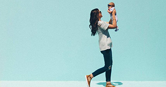 A woman wearing jeans and striped t-shirt smiles and holds a small baby in the air while walking.