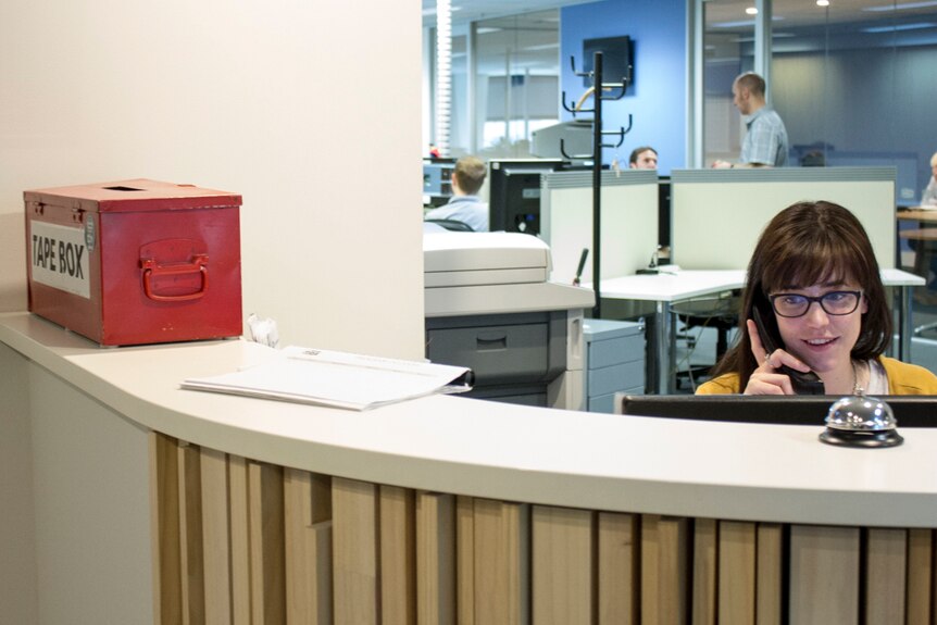 A woman sits at a reception desk. People working in the background.