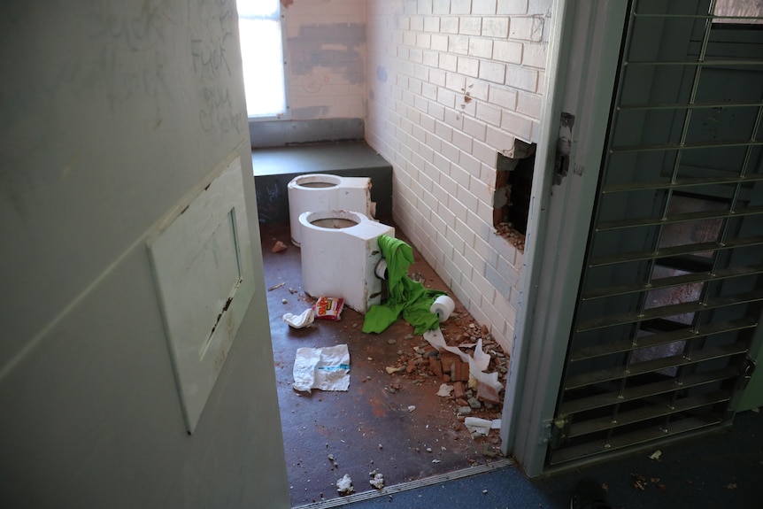 Toilets have been ripped out of walls in a damaged prison cell seen through an ajar door.