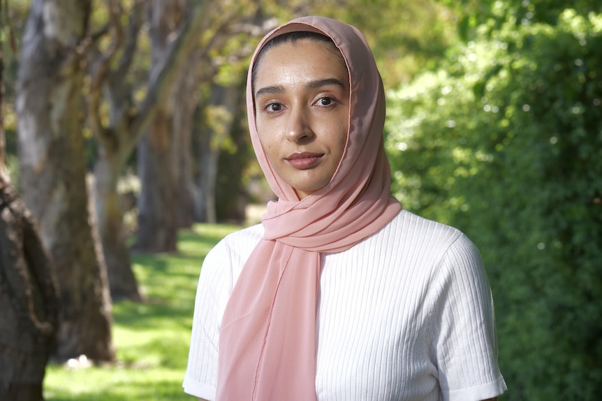 Woman in pink headscarf and white t-shirt stands outside, with trees in background.
