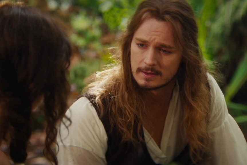 A man with long-hair sits in a garden talking to a woman. His face looks like it's been added by CGI.