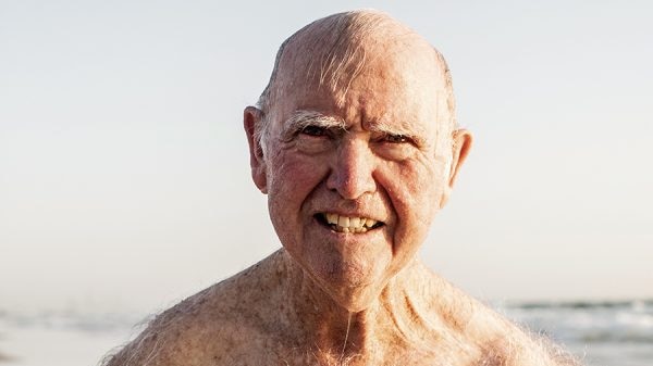older man in togs at the beach smiling at the camera