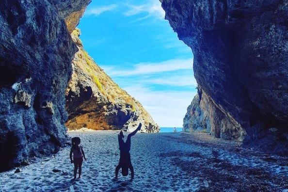 one child standing and another doing a handstand inside a cave with the beach behind