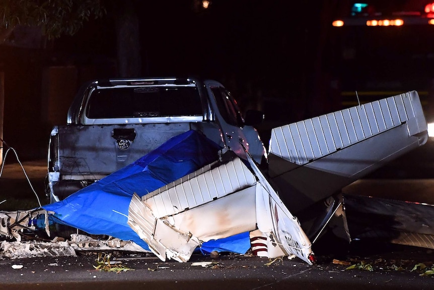 The burnt out wreckage of a light plane sits smashed in the middle of a street with a wing pointed to the sky.