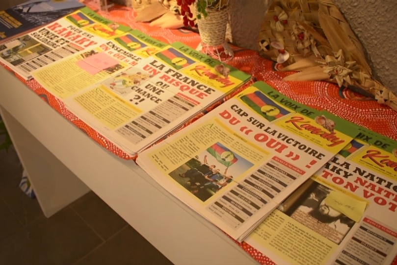 Under yellow lamp light, you view a tabled filled with the same newsletters that have 'La voix de Kanaky' as its title.