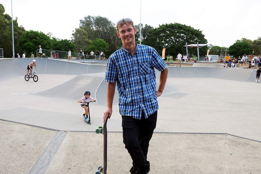 Man with blue and white check shirt leans on a skateboard in front of the skate park