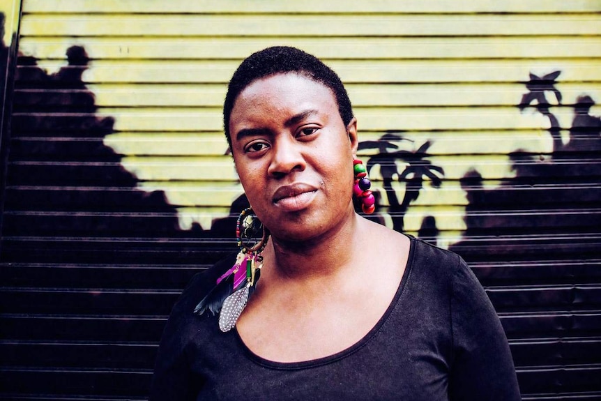 The writer stands against a corrugated garage door  painted with a yellow and black mural. She wears dream-catcher earrings.