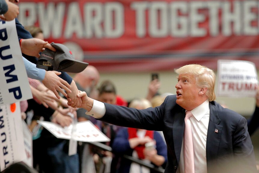 Side on view of Donald Trump reaching up to shake hands with supporters, only their arms visible.
