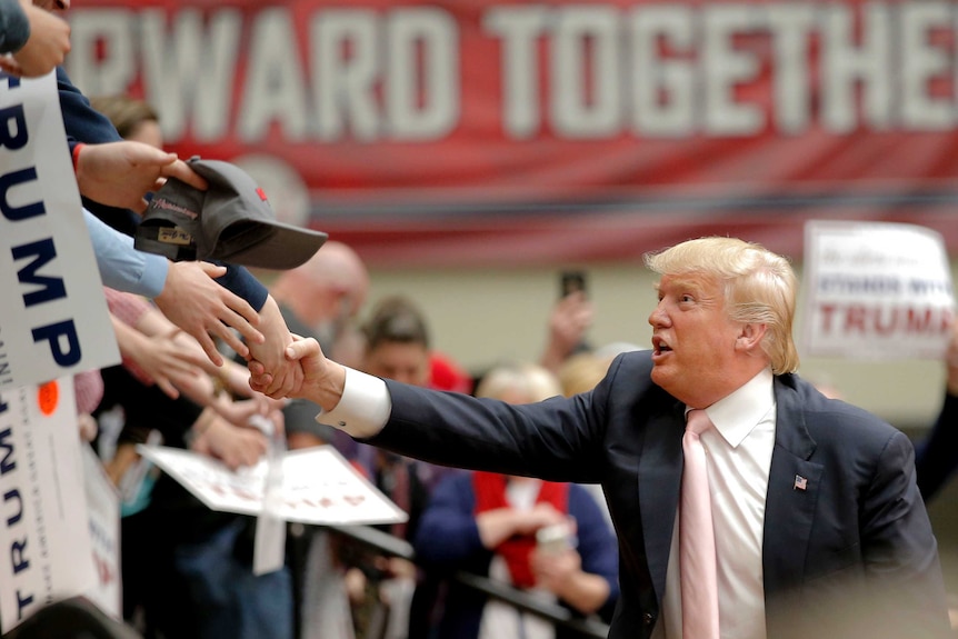Side on view of Donald Trump reaching up to shake hands with supporters, only their arms visible.