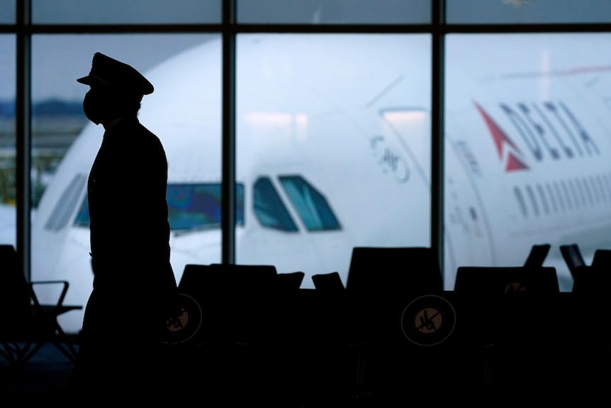 A silhouette of a pilot on a mask walking through an airport with a Delta airplane seen out the window behind. 