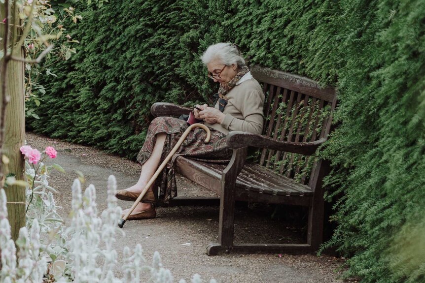 An elderly woman sitting on a bench in a garden with a walking stick lying against her leg
