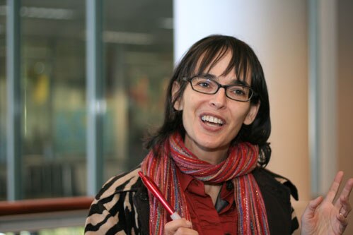 Woman with dark hair and dark glasses and red scarf talks animatedly to the camera