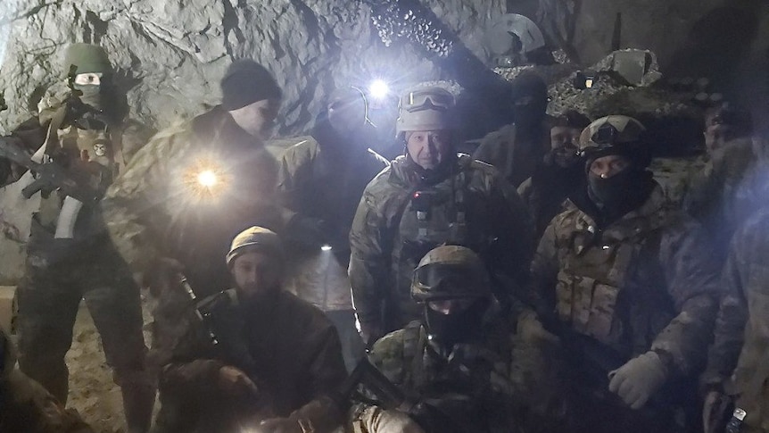 A group of men in combat fatigues holding weapons and torches pose in front of a rocky, white, underground wall.
