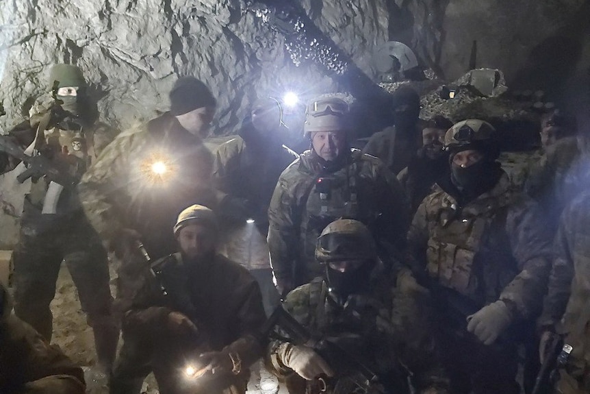 A group of men in combat fatigues holding weapons and torches pose in front of a rocky, white, underground wall.