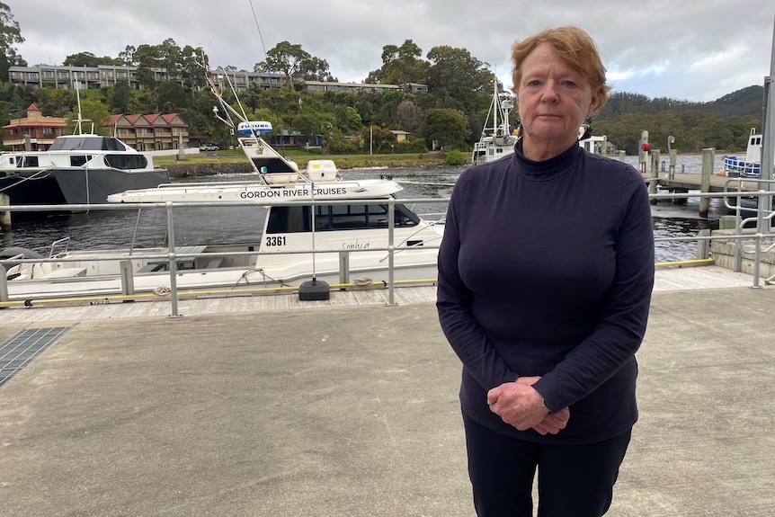 A woman wearing a dark shirt stands on a concrete harbour in front of boats.