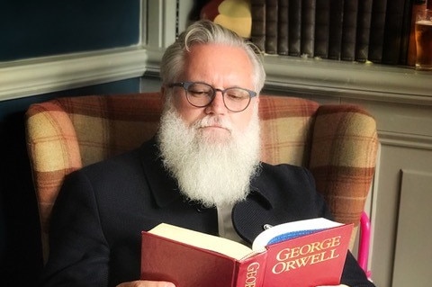 A man sits in an armchair reading a book on George Orwell.