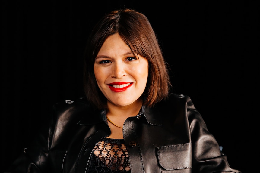 Nakkiah wearing a black top sitting on a chair smiling 