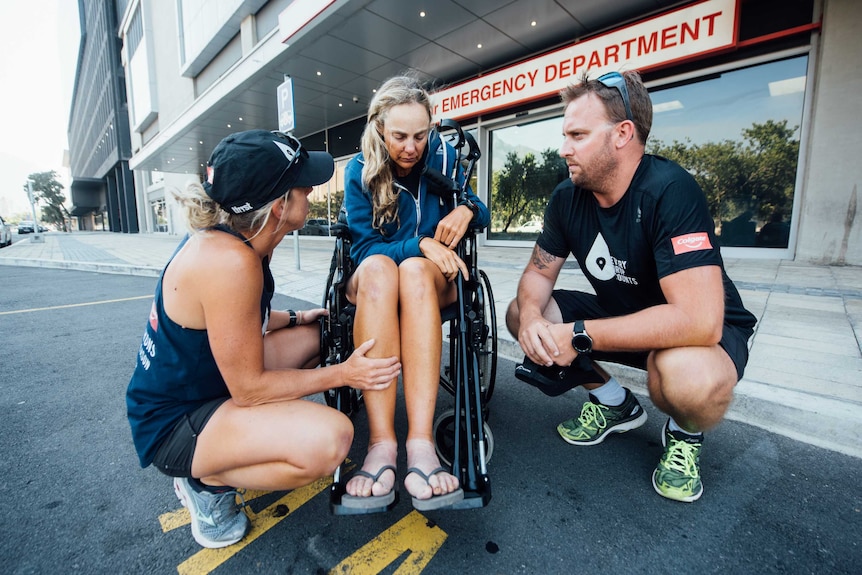 A woman looks tired and sad sitting in a wheelchair, consoled by a man and a woman outside an emergency department