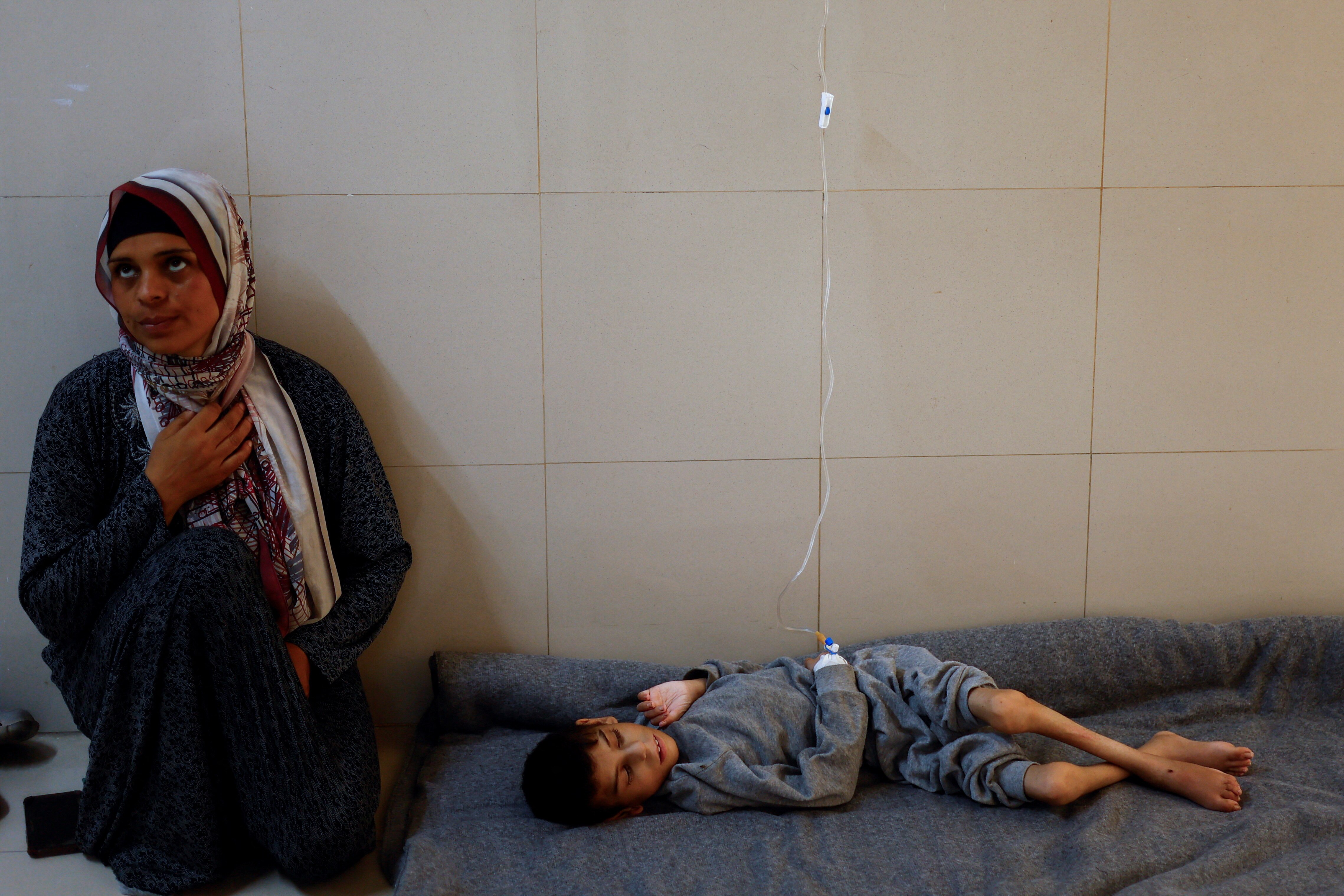A woman wearing a hijab sits on the hospital floor, holding hand over heart as her son lies on a blanket connected to an IV drip