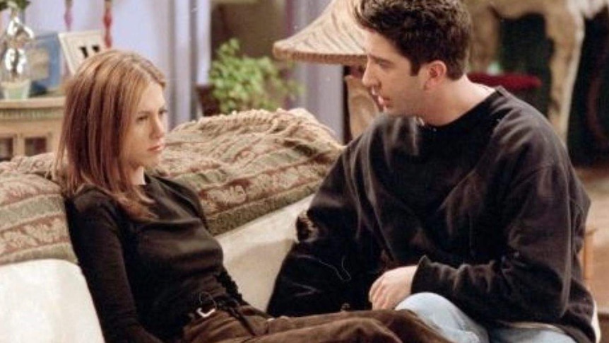 Rachel from the TV show friends sits on a couch and looks away from her boyfriend Ross who sits next to her trying to talk.