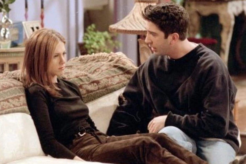 Rachel from the TV show friends sits on a couch and looks away from her boyfriend Ross who sits next to her trying to talk.