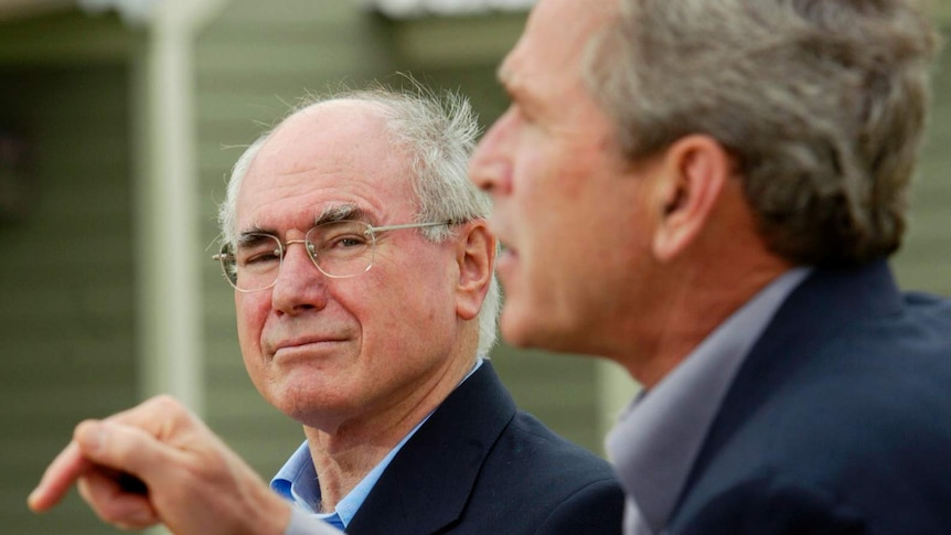 a man in glasses watches as a man points and speaks at a microphone