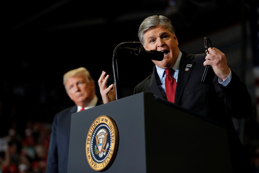 Sean Hannity speaks at a podium at a Trump rally, with Donald Trump in the background. 