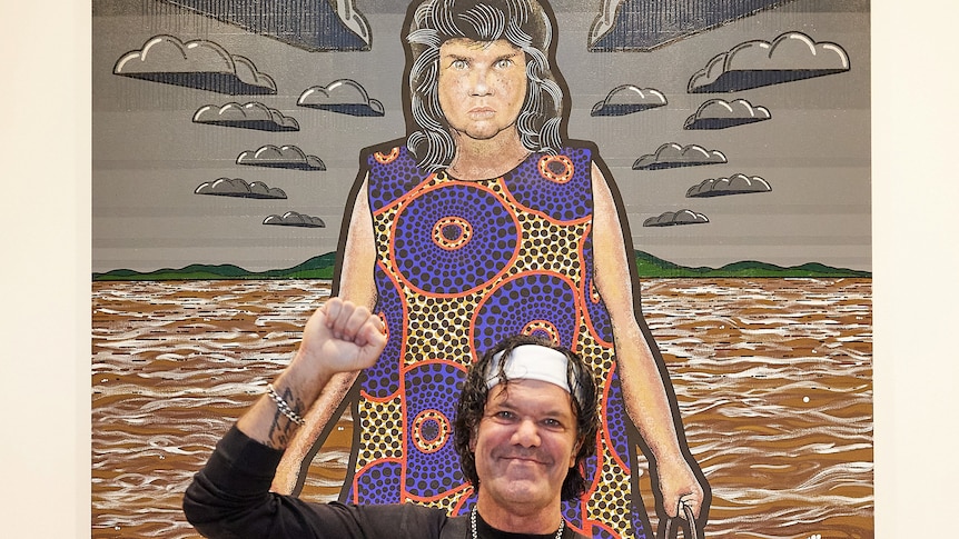 An Indigenous man smiles, one fist raised in celebration, while standing in front of a large portrait of an Indigenous woman
