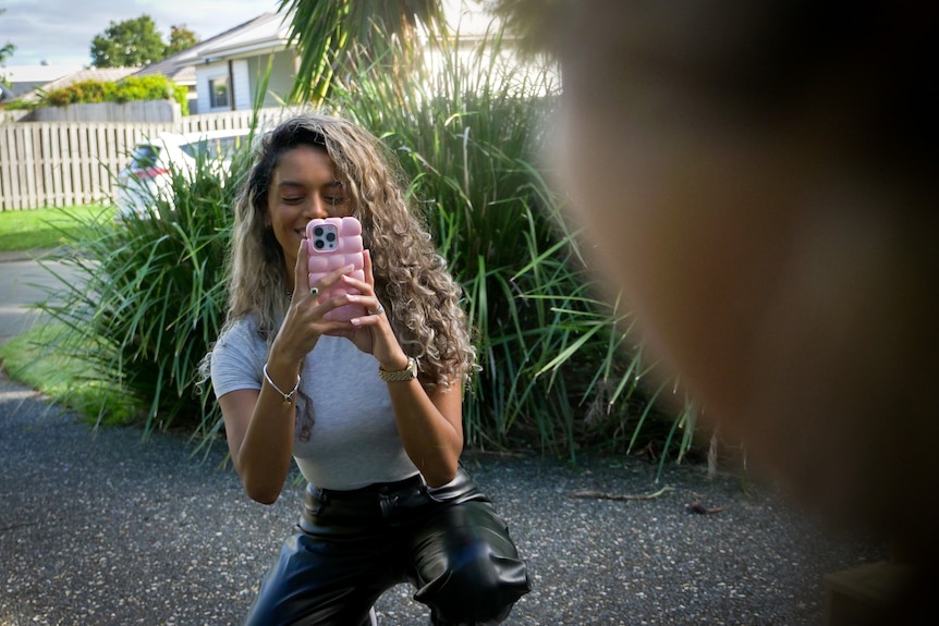 A woman crouches on a driveway, holding up an iPhone in a pink case to take a photo.